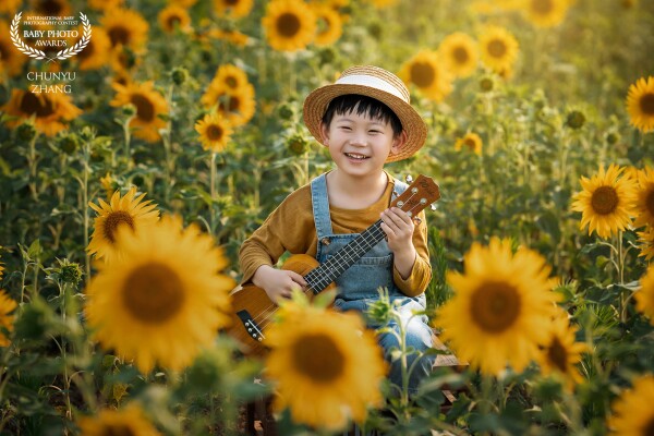 This little boy is particularly fond of music. One day, he came to a beautiful field of sunflowers and started to play the ukulele. The weather was fine, the sunflowers were blooming brilliantly, and he had a great afternoon.