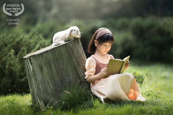 This little girl is sitting on the grass reading a book. Look, who is quietly climbing behind her? A little rabbit sneaks up on a tree stump and is also reading her book.