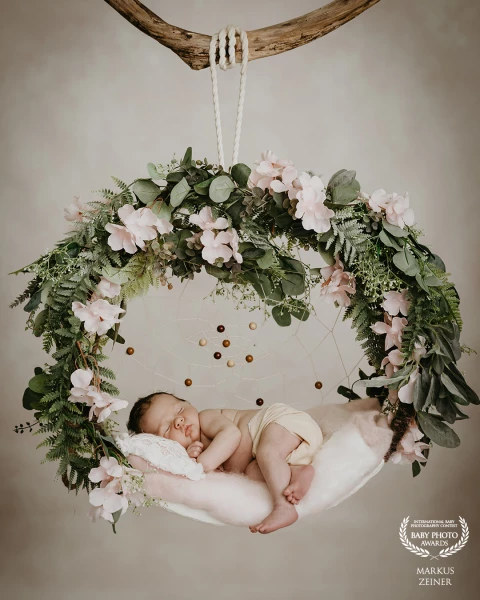 A sleeping newborn rests on a soft hanging swing, nestled within a dreamy floral dreamcatcher of euc...