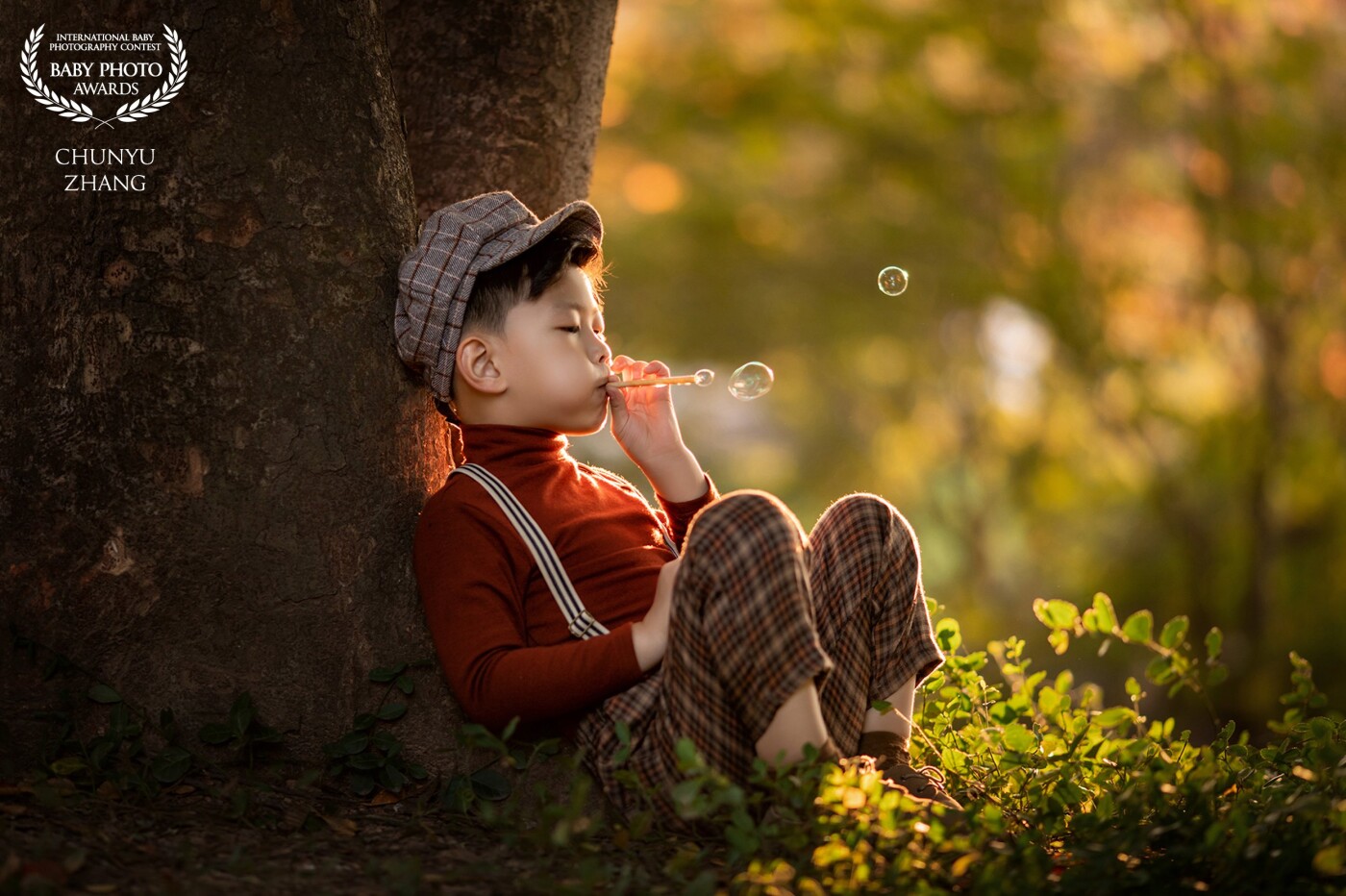 This little boy is playing in the sunset, he is leaning against a big tree. He is blowing bubbles. He really enjoys this good time playing all by himself.