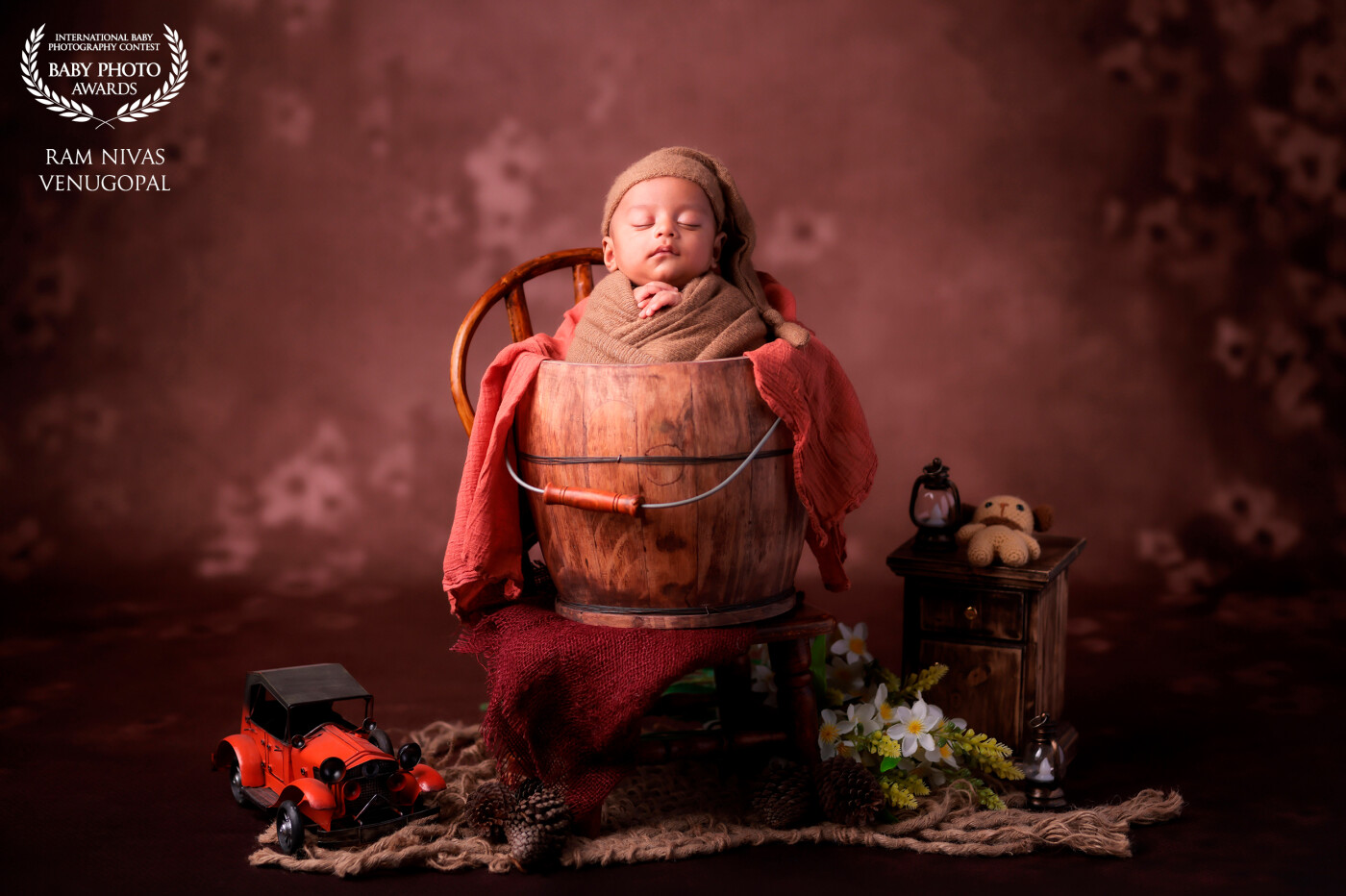 In a chiaroscuro of innocence and tranquility, this photograph showcases a sleeping newborn, delicately wrapped and cradled in a bowl on a chair. The image is enhanced by the playful interplay of shadows and light, highlighting the baby's peaceful features against a dark backdrop. Flanked by whimsical props including a small car, a lantern, flowers, and a woven toy, the composition creates a serene tableau, beautifully juxtaposing the vibrant infancy against the quietude of the setting.