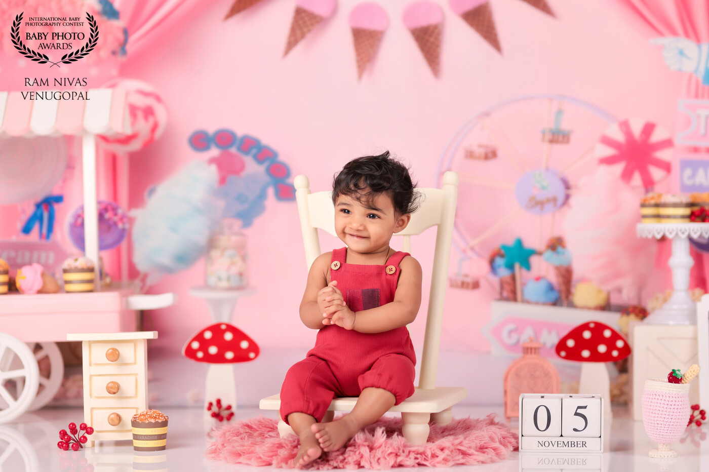 In this delightful baby photography image, a charming little boy sits in a white chair, his face adorned with a captivating smile that fills the frame with wonder. The backdrop is reminiscent of a whimsical fun land, transporting us to a world of enchantment. The scene is adorned with delightful props, including a candy shop and an array of delectable cakes and desserts, adding a touch of sweetness to the captivating composition.