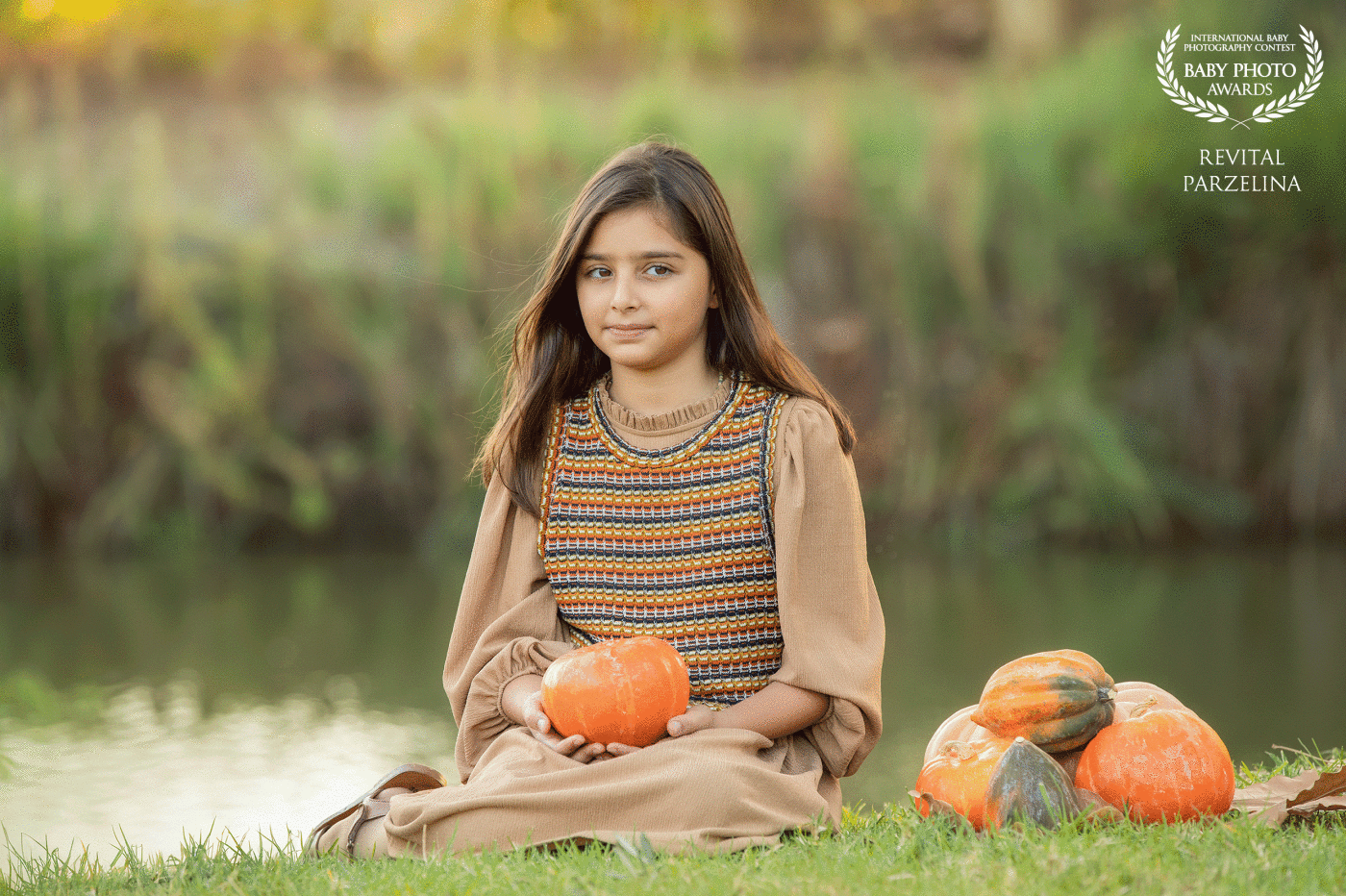 "Captured in the serene embrace of nature, a young girl sits gracefully on the lush green grass, cradling a pumpkin in her hands. The gentle breeze whispers through the nearby trees as the tranquil river flows beside her, creating a harmonious scene that reflects the beauty of the autumn season. The girl's connection to the earth and the simplicity of the moment are encapsulated in this enchanting photograph, where the vibrant orange hues of the pumpkin complement the natural surroundings, inviting a sense of warmth and tranquility."