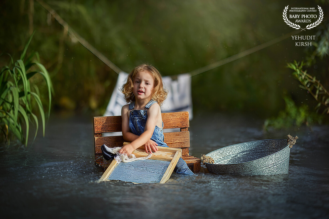 "River Days" <br />
With a nostalgic nod to simpler times, this enchanting portrait captures a little girl immersed in imaginative play, seated mid-river with a vintage washboard and denim overalls. Sunlight dances on the water around her, while a soft backdrop of green foliage and a clothesline of freshly washed linens sets the scene. Yehudit Kirsh masterfully blends storytelling and fine art, evoking innocence, freedom, and the boundless creativity of childhood. This image is both a tribute to natureโs quiet beauty and to the whimsical world of a childโs imagination.