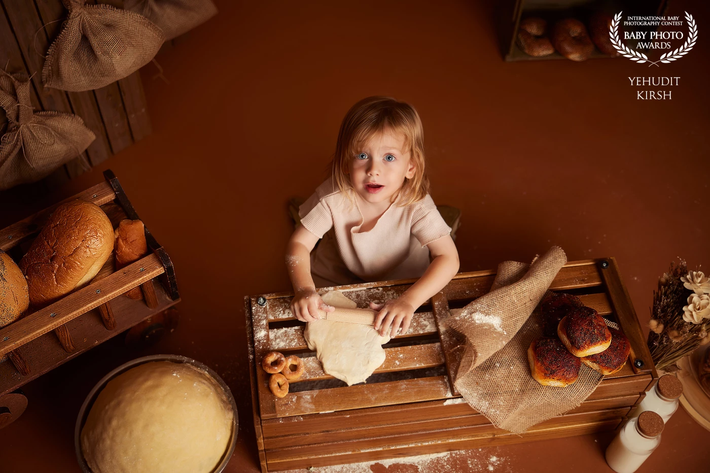 In a warm, rustic kitchen scene, a young boy on a wooden crate, fully immersed in rolling out dough with both hands. His blonde curls frame a focused, curious expression as he explores the simple joy of baking. Thereโs no apron or hat โ just a natural, authentic moment of childhood, filled with charm and sincerity.<br />
<br />
Surrounded by traditional baked goods like challah, pretzels, and loaves of bread, the setting feels timeless, blending heritage with imagination. Though not a professional bakery, the scene radiates the essence of tradition, play, and creativity. cultural texture, and the beauty of everyday magic.