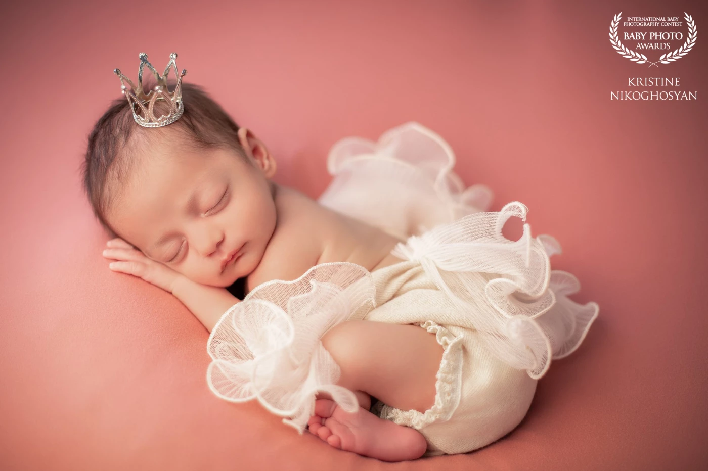 Cradled in the hush of rosy light, this little angel rests in a whisper of ruffles, crowned with the quiet grace of new life.