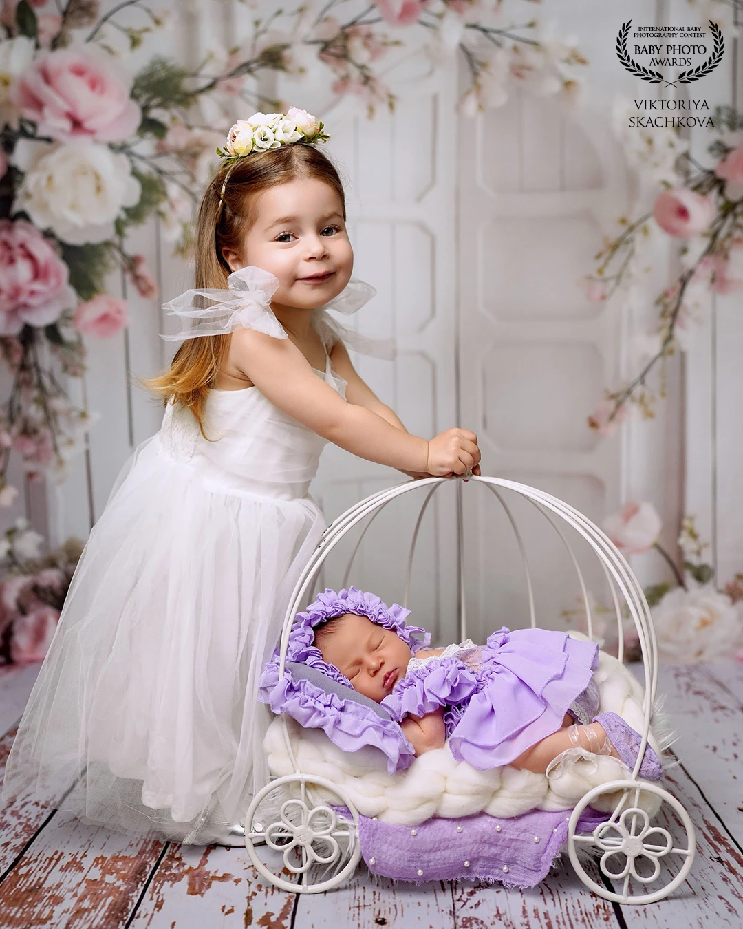 A gentle moment between sisters — the beginning of a lifelong bond. The proud big sister, dressed like a little angel, lovingly watches over her newborn sibling resting peacefully in a delicate carriage. Surrounded by soft florals and pastel tones, this image captures the beauty of family, tenderness, and the magic of new beginnings.