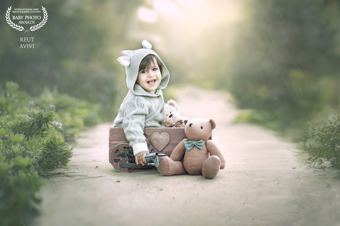 A one-year-old boy smiles sweetly in clothes adapted for him, happily sitting inside a wooden cart while leaving his teddy bear outside, adding a charming touch to the photo.