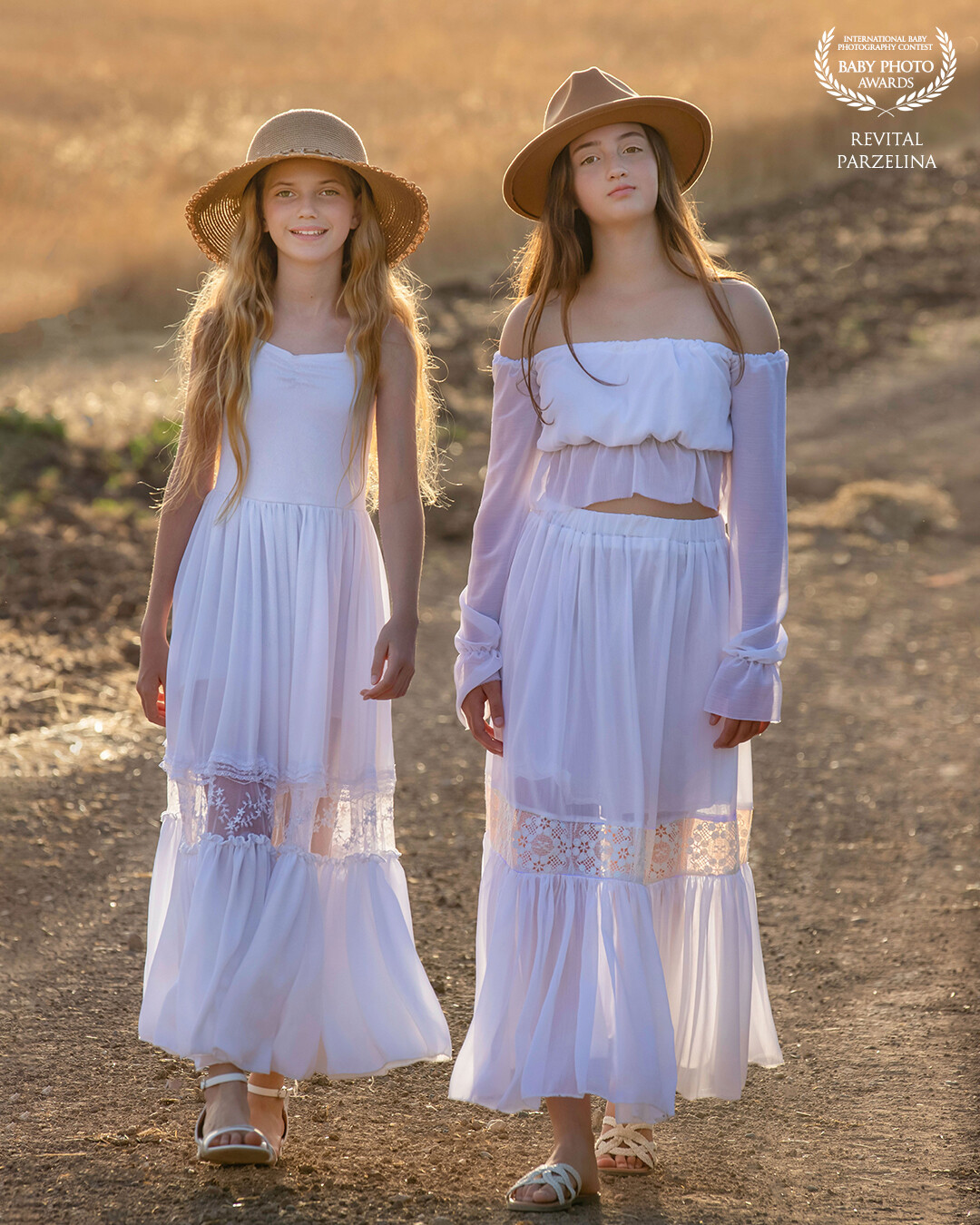 In this beautiful picture we can see a vast golden wheat field stretching endlessly in the background, while two lovely young girls stand in the foreground. Both girls wear a long white dress and a classic wide-brimmed straw hat, which perfectly complement the rural landscape. The sunlight casts a warm, glowing light on their playful faces, and their cheerful expressions radiate pure happiness and contentment. The girls are no doubt enjoying the fresh air and freedom of the open fields in this delightful moment of peace and quiet.